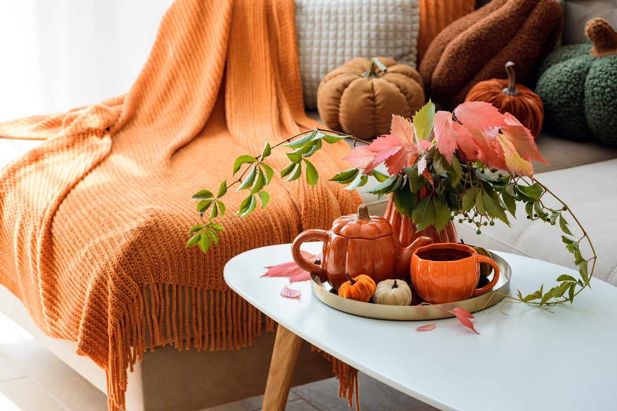 Warm autumn still life with decorative pumpkins, a knitted orange throw draped over a sofa, and a tray with a teapot and mug